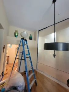 A room under renovation featuring a ladder decorative vases and a large pendant light. Walls are taped for painting